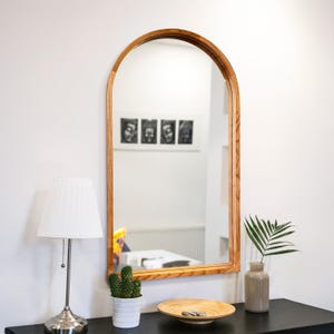May include: A wooden-framed arched mirror hangs on a white wall. The mirror reflects a room with artwork and furniture. A lamp, cactus, wooden bowl, and vase with a plant sit on a black table below the mirror.