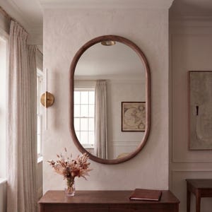 May include: An oval mirror with a dark wood frame mounted on a textured off-white wall above a rectangular wooden console table. The mirror reflects a room with a window, curtains, and a framed map. A brass ceiling light is positioned above.