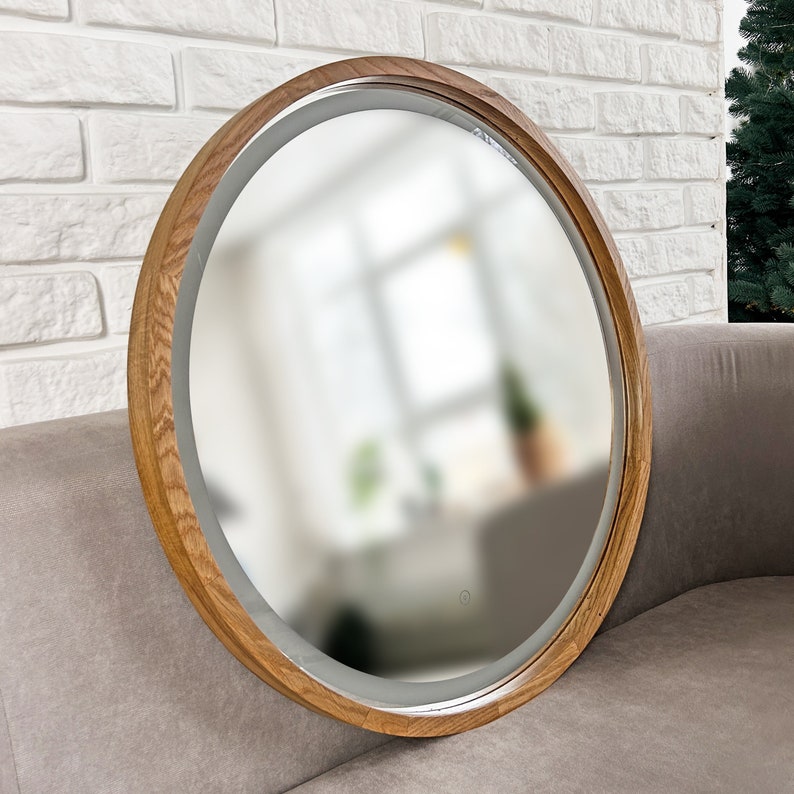 May include: A round wall mirror featuring a thick, natural-toned wooden frame with a visible grain pattern. The mirror includes an integrated LED light strip along the inner edge and a small, circular touch-sensitive power button on the glass surface.