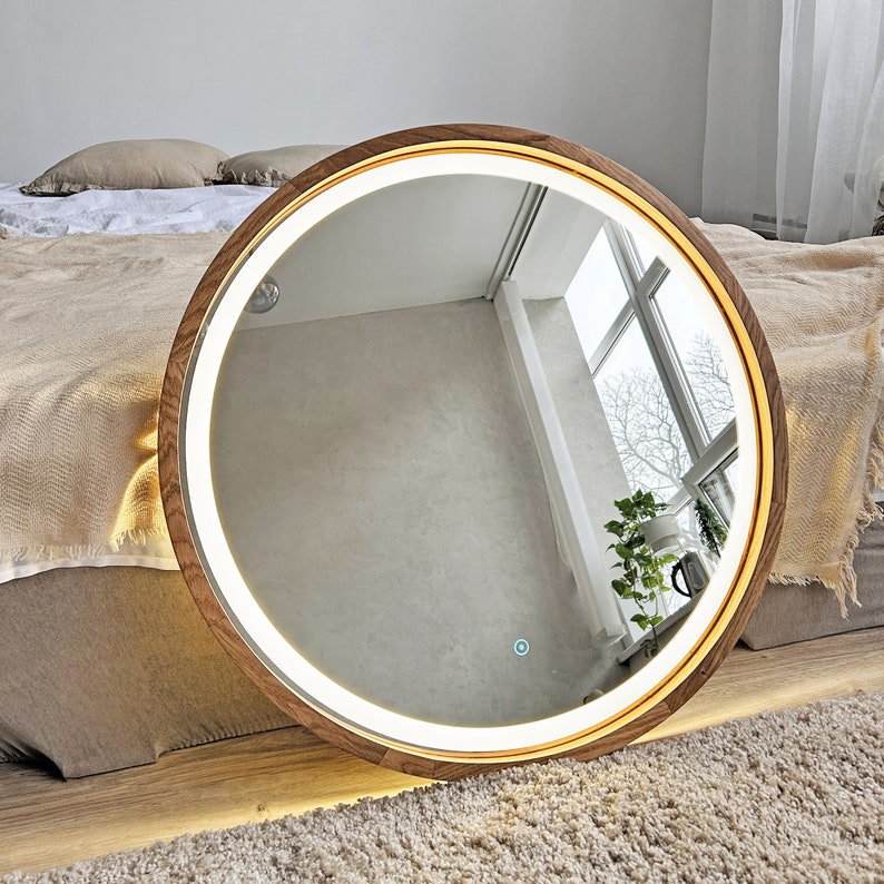 May include: A circular wall mirror featuring a warm-toned wooden frame and an integrated LED light strip that illuminates the inner edge. The mirror surface includes a small, glowing touch-sensitive power button at the bottom center.