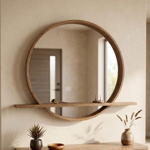 Op de afbeelding: Een ronde wandspiegel met een frame van natuurlijk hout en een geïntegreerde horizontale plank die door het midden loopt. De spiegel reflecteert een deuropening en een binnenruimte, terwijl op de plank een kleine metalen sleutelbos ligt.