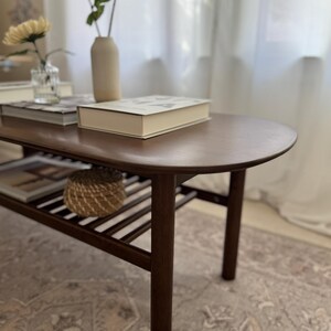 Puede incluir: Una mesa de centro de madera marr&oacute;n oscuro con una parte superior redondeada y un estante inferior. La mesa tiene un acabado de madera natural y est&aacute; sobre una alfombra estampada.