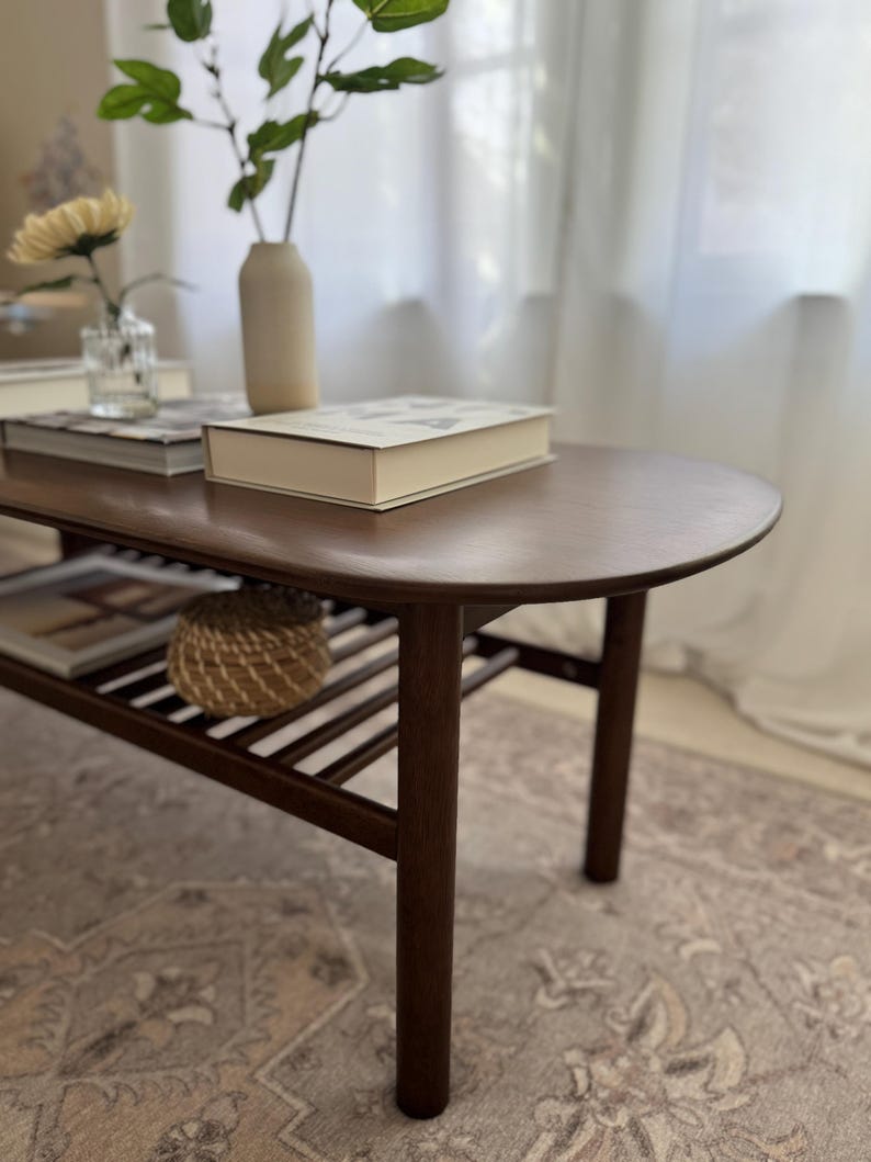 Puede incluir: Una mesa de centro de madera marr&oacute;n oscuro con una parte superior redondeada y un estante inferior. La mesa tiene un acabado de madera natural y est&aacute; sobre una alfombra estampada.