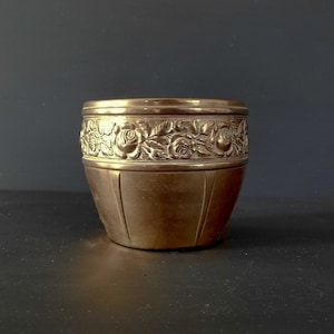 Könnte beinhalten: Ein goldfarbener Metalltopf mit einem dekorativen Rosenmuster am oberen Rand. Der Topf hat eine leicht abgerundete Form und eine glatte Oberfläche.