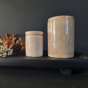 Puede incluir: Dos tarros de cerámica, uno alto y otro bajo, ambos de color beige claro y con una superficie lisa ligeramente texturizada. Los tarros están sobre una superficie negra.