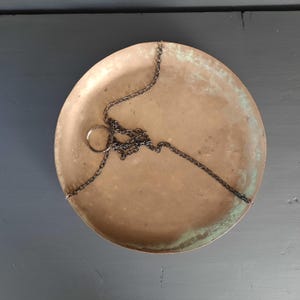 May include: A circular brass scale pan with a dark metal chain, showing signs of age and wear. The pan has a ring for hanging and a weathered, tarnished finish. The overall style is vintage and rustic, with a hint of green.