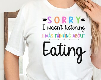 May include: White t-shirt with the text "SORRY I wasn't listening I WAS THINKING ABOUT Eating" in colorful letters.