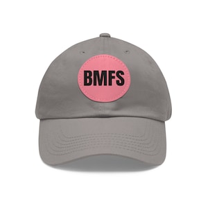 Può includere: Un cappellino da baseball grigio con visiera curva. Una toppa circolare rosa sul davanti mostra le lettere nere "BMFS". Il cappello è realizzato in un materiale morbido e ha un cinturino regolabile.
