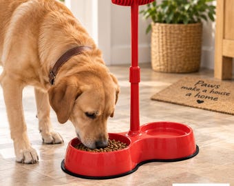 May include: A bright red plastic pet feeder with two circular bowls and a central vertical handle with a ribbed grip. The feeder is positioned on a tiled floor, and a golden-coloured dog is eating dry kibble from one of the two red bowls.