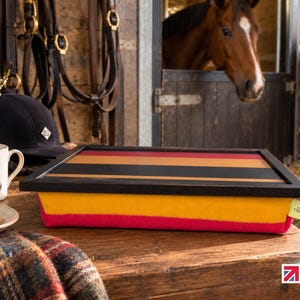 May include: A lap tray with a black frame and a surface featuring horizontal stripes in red, black, and tan. The base is a soft, bright yellow and pink cushion. A small tag on the side reads Made in the Mill. The tray sits on a wooden surface.
