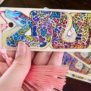 May include: A colorful bookmark featuring a detailed illustration of a snake with a pink tassel. The snake is depicted in a vibrant, psychedelic style with a mix of blue, green, yellow, pink, and purple colors.