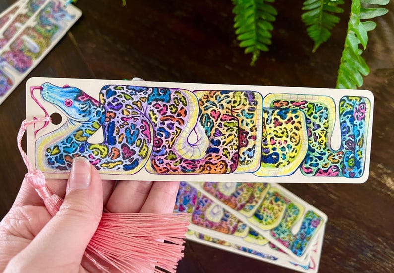 May include: A colourful bookmark featuring a detailed illustration of a snake with a pink tassel. The snake is depicted in a vibrant, psychedelic style with a mix of blue, green, yellow, pink, and purple colours.