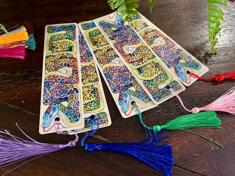 May include: Four colourful bookmarks featuring a detailed illustration of a snake with a patterned skin. Each bookmark has a different coloured tassel.