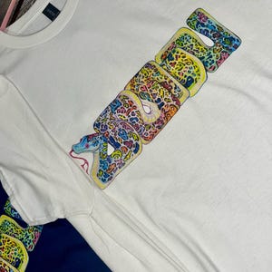 Peut inclure: T-shirt blanc avec un motif de serpent coloré. Le mot "SNAKE" est épelé en grandes lettres stylisées, chaque lettre présentant un motif unique de couleurs vives et d'imprimés animaliers. La tête du serpent est au début du mot.