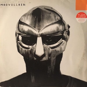 May include: A black and white photo of a person wearing a silver metal mask. The mask has a large, prominent nose and a wide, flat forehead. The person's eyes are visible through the mask, and they are looking directly at the camera. The text "MF DOOM & MADLIB PRESENT MADVILLAIN" is visible in the upper right corner of the image.