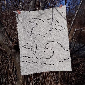 May include: A white crocheted blanket with a geometric design. The blanket is hanging on a tree branch.