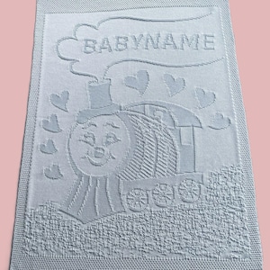 May include: A light blue knitted baby blanket with a white embroidered design of a train with a smiling face and the words "BABYNAME".