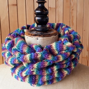 Könnte beinhalten: Handgestrickter Loop-Schal in leuchtenden Farben. Er zeigt eine Mischung aus Lila, Blau, Grün und Creme. Der Schal mit grobem Strick wird auf einer Vintage-Büste präsentiert, die sein strukturiertes Design und seine gemütliche Ausstrahlung zeigt. Ein stilvolles Accessoire.