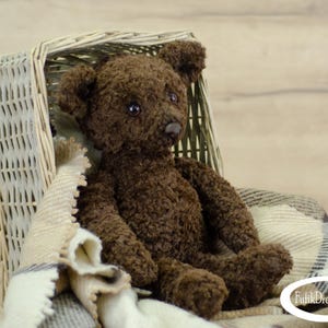 May include: A brown teddy bear sitting in a wicker basket lined with a cream and gray plaid blanket. The bear has black eyes and a black nose.