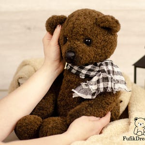 May include: A handmade brown teddy bear with a black and white checkered scarf. The bear has a textured fur appearance and is being held by a person. The bear is sitting on a cream-colored blanket.