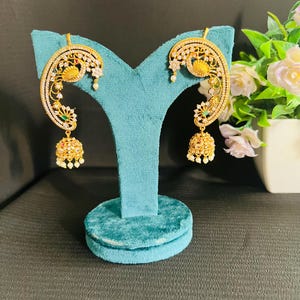 May include: Gold-toned earrings with a peacock design, featuring white gemstones and dangling gold bells. The earrings are displayed on a teal velvet stand, with a floral arrangement in the background.