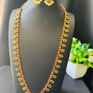 May include: A gold-toned necklace and matching earring set featuring teardrop-shaped links with embedded green stones and small pink accents. The necklace includes a central pendant with a green stone and dangling white beads.