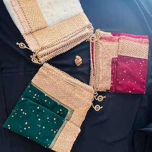May include: Three decorative fabric pieces in white, burgundy, and emerald green, each with gold trim and embellishments. The white piece has pearl-like accents. A small gold-colored figurine is centered between the fabrics. The fabrics are folded and arranged on a dark surface.