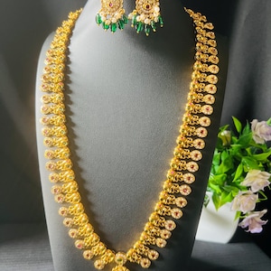 May include: A gold-toned necklace and matching earring set featuring intricate metalwork, small embedded red and white stones, and dangling green teardrop-shaped beads. The long necklace has a central pendant with similar stone and bead accents.