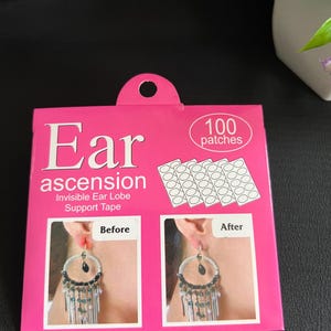 May include: A pink package of "Ear Ascension" invisible ear lobe support tape. The package shows a before and after image of earrings. The package also includes 100 patches. The text on the package reads "Ear ascension Invisible Ear Lobe Support Tape".