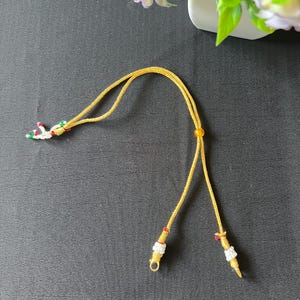 May include: A golden yellow cord necklace featuring a sliding bead adjustment mechanism. The ends of the cord are decorated with small white, red, and green beads, while a single amber-colored bead is positioned along the length of the cord.