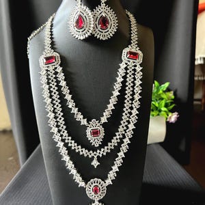 May include: A jewelry set consisting of a multi-layered necklace and matching drop earrings. The pieces are crafted from silver-toned metal with clear white stones and deep red rectangular and pear-shaped center stones in a floral design.