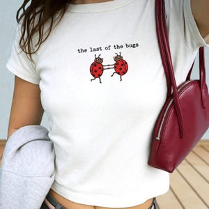 May include: A cream-coloured short-sleeve t-shirt featuring a graphic of two red ladybirds holding hands with the text "the last of the bugs" printed above them in a black typewriter-style font, paired with a dark red shoulder bag.