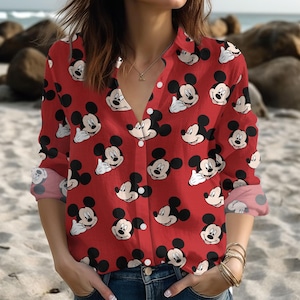 Puede incluir: Una camisa roja con botones con un patrón repetido de caras de Mickey Mouse. La camisa de manga larga tiene un cuello y puños clásicos, con un escote en V. Los puños están doblados, revelando un patrón de lunares rosas.