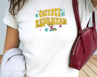 Może przedstawiać: Biały t-shirt z krótkim rękawem z żółtym napisem OUTFIT REPEATER w czcionce bąbelkowej, któremu towarzyszy mała postać z kreskówki, różowe gwiazdki i niebieskie kwiaty. Ciemnoczerwona torba na ramię ze srebrnym zamkiem błyskawicznym.