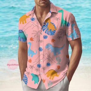 Op de afbeelding: Roze shirt met korte mouwen met een dinosauruspatroon. Het shirt heeft een relaxte pasvorm en is versierd met kleurrijke dinosaurusillustraties, palmbomen en berggraphics. De achtergrond is een lichtblauwe oceaan.