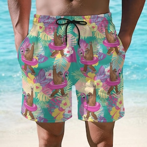 May include: Swim trunks with a vibrant pattern featuring a cartoon Bigfoot character wearing sunglasses and a pink flamingo float. The design includes tropical leaves and flowers in shades of pink, yellow, and green. The shorts have a drawstring waist.