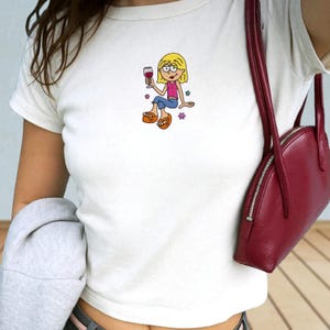 May include: A white short-sleeved t-shirt featuring a small graphic of a blonde cartoon character wearing a pink top and blue pants, holding a wine glass, with small colorful flower icons surrounding the figure, paired with a red shoulder bag.