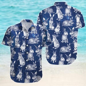 Puede incluir: Una camisa azul de manga corta con botones y un estampado de conejos blancos. La camisa tiene un cuello blanco y un bolsillo en el lado izquierdo.