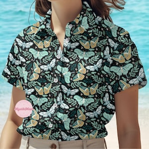 Puede incluir: Camisa negra con botones con un estampado repetido de polillas y hojas en tonos turquesa, dorado y blanco. La camisa tiene mangas cortas y un cuello clásico. El fondo es una imagen borrosa de una playa.