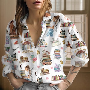 Pode incluir: Uma camisa branca de botões com mangas compridas apresenta uma estampa colorida de livros, flores e o texto "Compre livros para mim e me diga para ficar quieto".