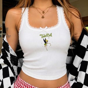 May include: A white ribbed vest top featuring a small graphic of Tinker Bell and the green text TinkerBell centred on the chest. The neckline and armholes are trimmed with white lace, and a small white ribbon bow is centred at the neckline.