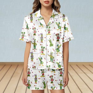 May include: White pajama set featuring a repeating pattern of cartoon characters including Mickey Mouse, Minnie Mouse, Donald Duck, and Goofy. The short-sleeved top and shorts have a button-down front and a contrasting trim.