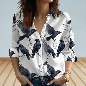 May include: White button-down shirt featuring a repeating pattern of black crows and leafy branches. The long-sleeved shirt has a V-neck and is made of a lightweight fabric. The crows are depicted in various poses, some with wings spread.