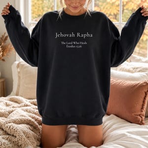 May include: A black long-sleeve crewneck sweatshirt featuring white text printed on the chest that reads Jehovah Rapha, The Lord Who Heals, and Exodus 15:26. The garment has a ribbed collar, cuffs, and hem with a relaxed fit.