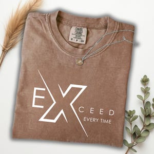 May include: A brown Comfort Colors t-shirt with the word "EXCEED" in white, along with the phrase "EVERY TIME". A silver necklace with a pendant rests on the shirt. The shirt is folded and displayed with decorative sprigs.