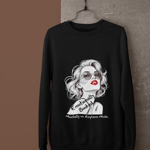 May include: Black sweatshirt featuring a stylized illustration of a woman with sunglasses and red lipstick. The text reads "Emotionally Booked, Mentally on Airplane Mode."