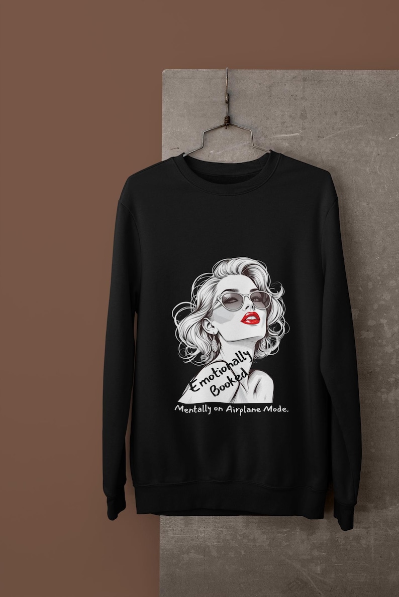 May include: Black sweatshirt featuring a stylized illustration of a woman with sunglasses and red lipstick. The text reads "Emotionally Booked, Mentally on Airplane Mode."