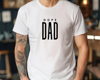 Peut inclure: T-shirt blanc avec l'inscription "DOPE DAD" en lettres noires et grasses. Le t-shirt a un col rond et des manches courtes. Le texte est centré sur la poitrine.