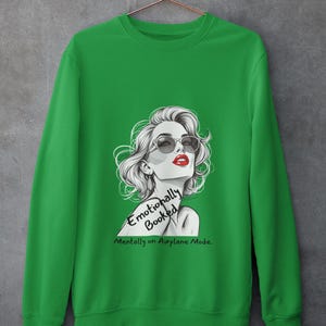 May include: Green sweatshirt featuring a graphic of a woman with sunglasses and red lipstick. The text on the sweatshirt reads "Emotionally Booked, Mentally on Airplane Mode."