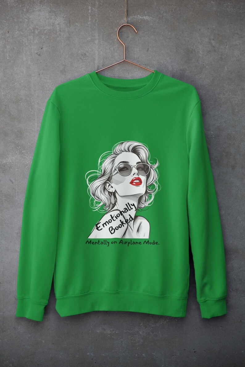 May include: Green sweatshirt featuring a graphic of a woman with sunglasses and red lipstick. The text on the sweatshirt reads "Emotionally Booked, Mentally on Airplane Mode."